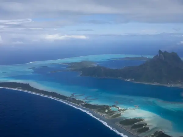 Lagoon and barrier reef encircling a volcanic island in the Pacific, with distant hills under clear sky