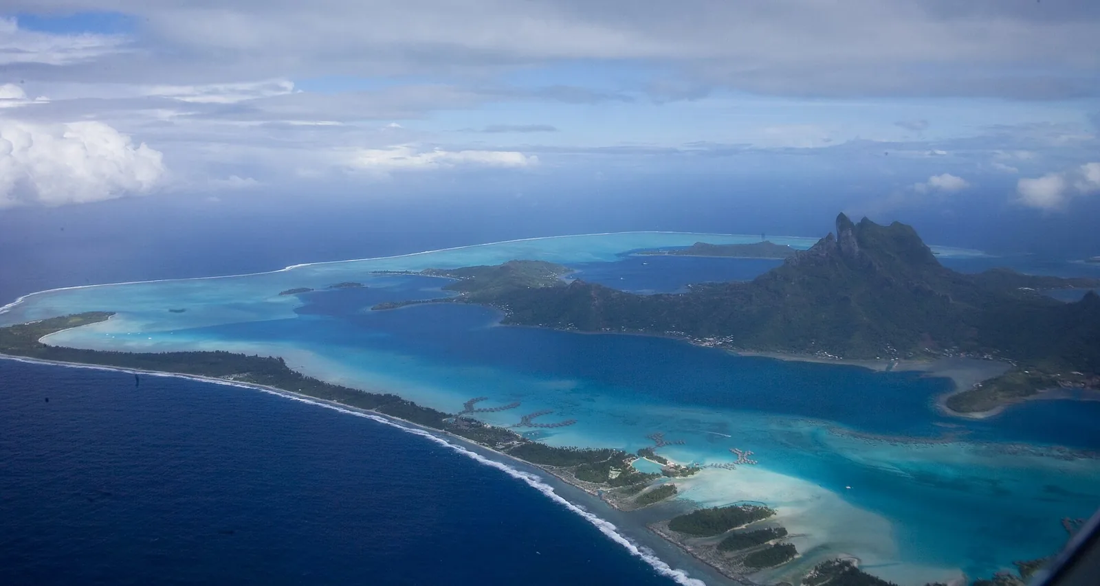 Lagoon and barrier reef encircling a volcanic island in the Pacific, with distant hills under clear sky